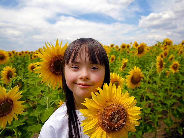 Brunette elementary aged girl with Down syndrome smiles in field of sunflowers on sunny day with clouds.