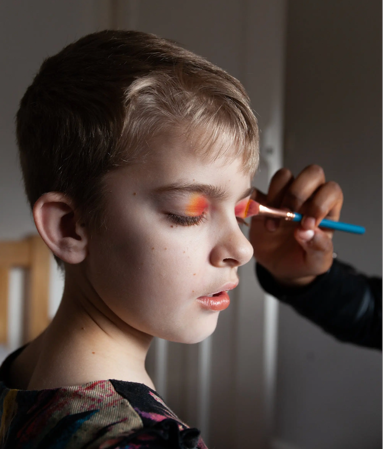 Closeup portrait of boy child getting orange eyeshadow applied, exploring gender roles and beauty standards.